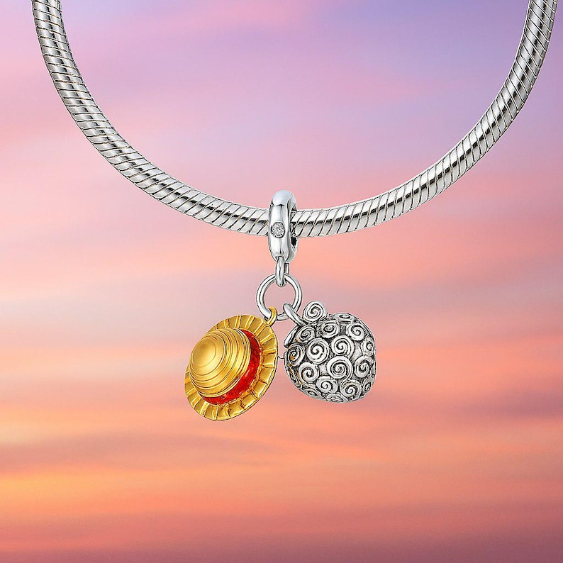 May include: A silver charm bracelet with two pendants. One pendant is a gold and red straw hat, and the other is a silver, patterned fruit. The background is a gradient of pink, orange, and purple.