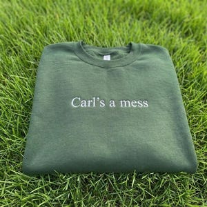 May include: A dark green sweatshirt with the embroidered text "Carl's a mess" in white. The sweatshirt is lying on a bed of green grass.
