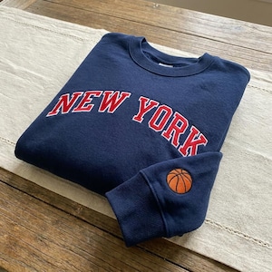 以下が含まれることがあります： 胸元に白い縁取りの赤いフォントでNEW YORKと刺繍されたネイビーブルーのクルーネックトレーナー。袖口付近には、黒いラインが入った小さな円形のオレンジ色のバスケットボールのグラフィックが刺繍されています。