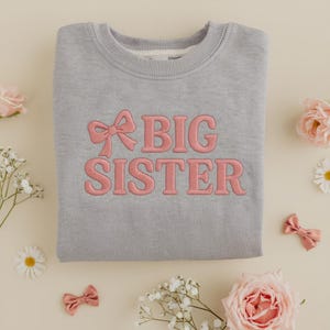 Könnte beinhalten: Ein graues Sweatshirt mit dem in Rosa gestickten Schriftzug "BIG SISTER". Über dem Wort "BIG" befindet sich eine rosa Schleife. Das Sweatshirt ist von Blumen und kleinen rosa Schleifen umgeben.