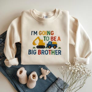 以下が含まれることがあります： クリーム色のスウェットシャツ。カラフルな文字で「I'M GOING TO BE A BIG BROTHER」と刺繍されています。デザインには黄色の掘削機と青いトラックが含まれています。スウェットシャツは、ベビーブーティー、テディベア、ジーンズと組み合わせています。
