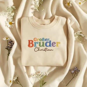 May include: Cream-colored sweatshirt with the embroidered text "Großer Bruder Christian" in colorful letters. The sweatshirt is surrounded by white flowers and rests on a cream-colored fabric.