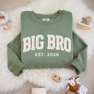 May include: A sage green long-sleeve sweatshirt featuring the text BIG BRO in large, cream-coloured block letters across the chest, with the text EST. 2026 printed in a smaller font directly underneath. The garment has a ribbed crew neckline and cuffs.