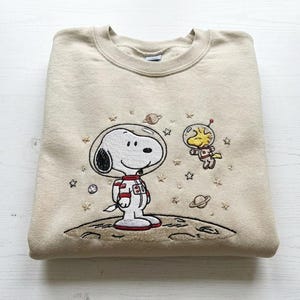Könnte beinhalten: Beiges Sweatshirt mit Rundhalsausschnitt und gesticktem Snoopy im Raumanzug, der auf einem Mond mit Sternen und Planeten steht. Ein gelber Woodstock im Raumanzug schwebt in der Nähe. Das Design ist in Weiß-, Rot-, Schwarz- und Gelbtönen gehalten.
