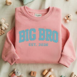 May include: A light pink long-sleeve sweatshirt featuring the text BIG BRO in light blue block letters across the chest, with the text EST. 2026 underneath in a smaller light blue font. The garment has a ribbed crew neckline and ribbed cuffs.