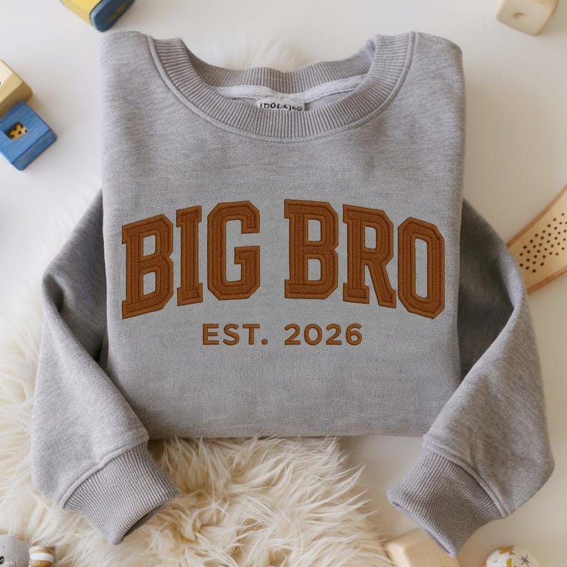 May include: A heather grey crewneck sweatshirt featuring the text BIG BRO in large, brown, block-style letters with a dark brown outline. Below the main text, the words EST. 2026 are embroidered in a matching brown font. The garment has ribbed cuffs.