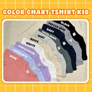 May include: A colour chart for kids' t-shirts displaying nine folded shirts in rose, pastel blue, purple pastel, white, beige, mint, muted green, navy, pepper gray, and black. Each shirt features a small white brand label at the inner neckline.