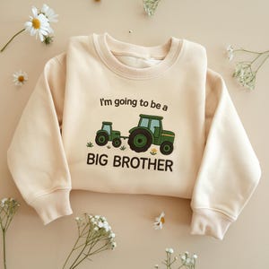 以下が含まれることがあります： 「I'm going to be a BIG BROTHER」の刺繍と緑色のトラクターのデザインが施されたクリーム色のスウェットシャツ。クルーネック、リブ編みの袖口と裾が特徴です。デザインには2台の緑色のトラクターと小さな花のアクセントが含まれています。