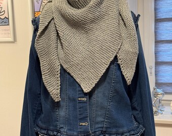 May include: A light gray knitted shawl draped over a dark blue denim jacket. The shawl has a triangular shape with a textured knit pattern. The jacket has a ruffled hem and button closures. The background includes framed artwork and a window.
