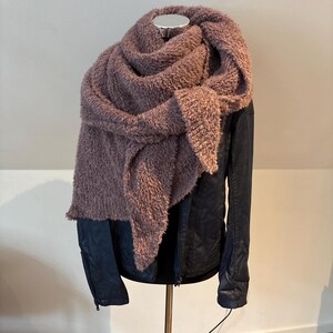 May include: A dusty rose-colored, fluffy scarf draped over a dark blue jacket. The scarf is thick and textured, creating a cozy and warm appearance. The jacket has a sleek, leather-like finish. The items are displayed on a mannequin.