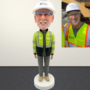 May include: A bobblehead figurine of a man wearing a white hard hat with the text "HPR ROB LUCCIONI". The figure is dressed in a green and black safety jacket, gray pants, and tan boots. A smaller image of the man in a safety vest is in the background.