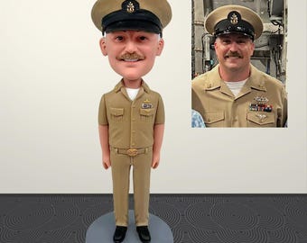 May include: A tan-colored bobblehead doll of a man in a U.S. Navy uniform, including a hat with a black brim. The doll is standing on a gray circular base. A smaller image of the man in uniform is in the background.