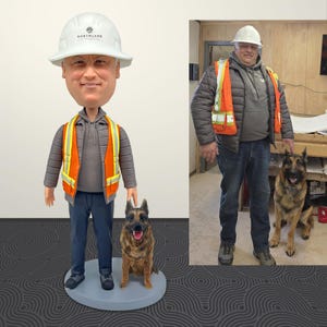 May include: A custom bobblehead of a man in a hard hat and safety vest, with a German Shepherd dog. The bobblehead has a gray base and the man's hard hat says "NORTHLAND". The image also shows a real man and dog.