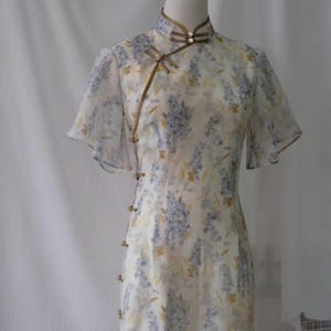 以下が含まれることがあります： 白い花柄のワンピース。チャイナカラーと短い袖が特徴です。青と黄色の花の繊細な模様があります。サイドにボタンがあり、襟と袖には茶色のトリムが付いています。
