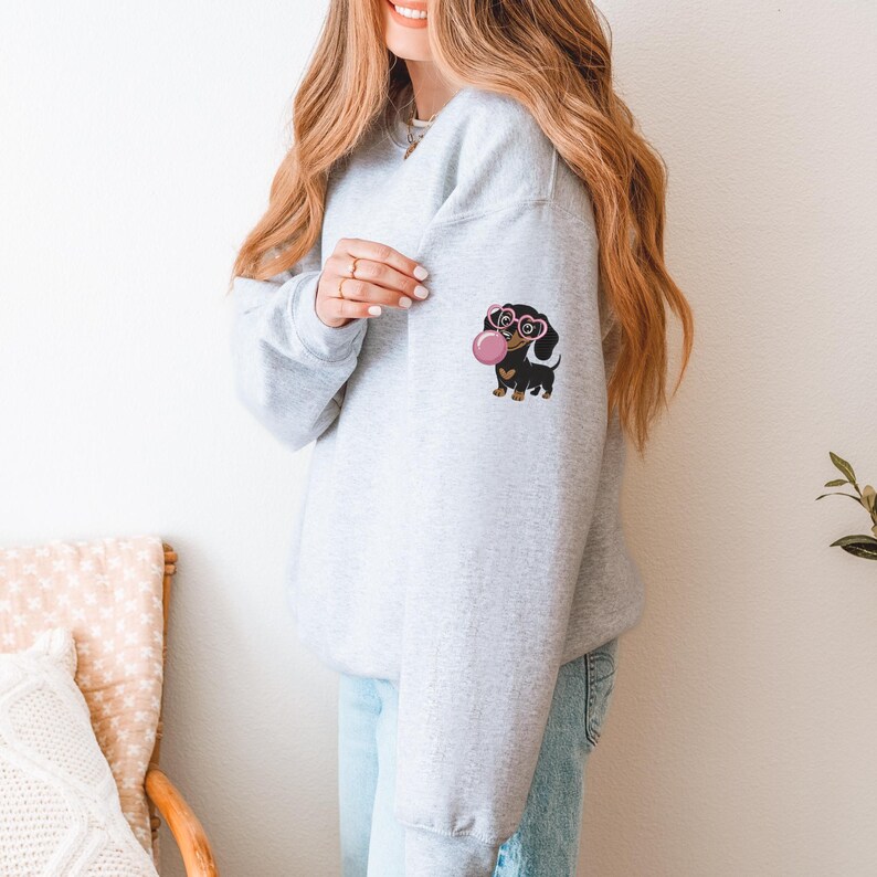 May include: Light gray sweatshirt featuring a dachshund graphic on the left sleeve. The dachshund is black and brown, wearing heart-shaped glasses and blowing a pink bubble. The sweatshirt is a casual, comfortable style.