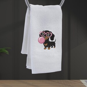May include: White hand towel with a cartoon dachshund wearing pink heart-shaped glasses and blowing a pink bubble. The dachshund has a brown chest and paws. The towel is hanging on a silver towel bar.