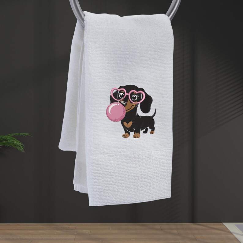 May include: White hand towel with a cartoon dachshund wearing pink heart-shaped glasses and blowing a pink bubble. The dachshund has a brown chest and paws. The towel is hanging on a silver towel bar.