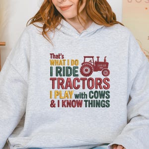Peut inclure: Un sweat-shirt gris clair avec l'inscription "That's What I Do, I Ride Tractors, I Play with Cows & I Know Things." Le motif comprend un tracteur rouge. Le texte est de couleurs vari&eacute;es.