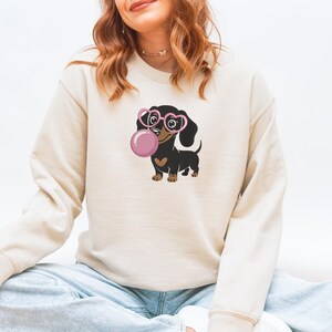 May include: Beige sweatshirt featuring a cartoon dachshund wearing pink heart-shaped glasses and blowing a pink bubble. The dachshund is black and brown with a heart-shaped patch. The sweatshirt is a neutral color.