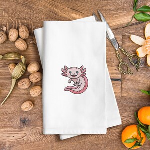 May include: White folded towel with an embroidered cartoon axolotl in pink and light pink. The axolotl has a smiling face and is centered on the towel. The towel is on a wooden surface with walnuts and scissors.
