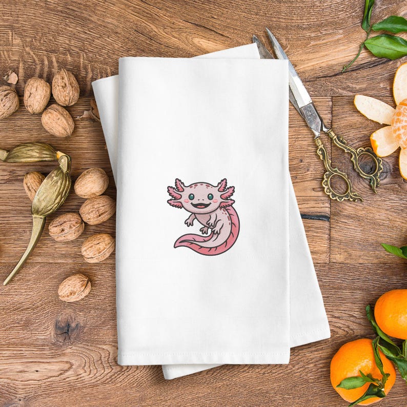 May include: White folded towel with an embroidered cartoon axolotl in pink and light pink. The axolotl has a smiling face and is centered on the towel. The towel is on a wooden surface with walnuts and scissors.