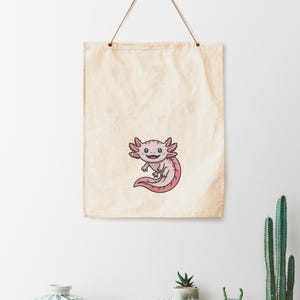 May include: A canvas wall hanging featuring a cartoon axolotl. The axolotl is pink with black outlines, a smiling face, and feathery gills. The background is a cream-colored canvas with a simple hanging cord.