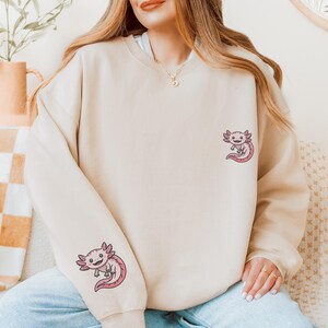 May include: Beige sweatshirt featuring two embroidered axolotl designs in pink and black. The axolotls are on the left sleeve and the upper right chest. The sweatshirt is a neutral color and has a crew neck.