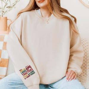 Peut inclure: Sweat-shirt beige avec le texte "That's what I do, I ride tractors, I play with cows & I know things" et un dessin de tracteur rouge sur la manche gauche. Le sweat-shirt est de couleur neutre et a un col rond.