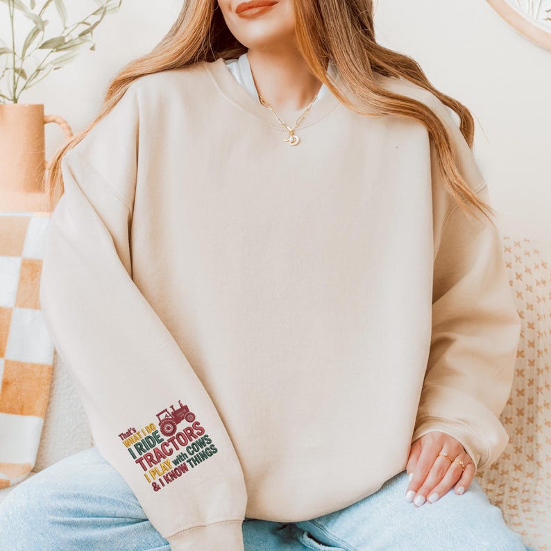 Peut inclure: Sweat-shirt beige avec le texte "That's what I do, I ride tractors, I play with cows & I know things" et un dessin de tracteur rouge sur la manche gauche. Le sweat-shirt est de couleur neutre et a un col rond.