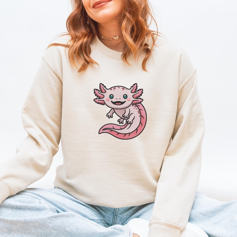 May include: Beige sweatshirt featuring a cartoon axolotl design. The axolotl is pink with black outlines and green eyes. The sweatshirt is a neutral color, suitable for casual wear.