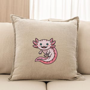 May include: Beige square throw pillow featuring an embroidered axolotl design. The axolotl is pink with black outlines, a smiling face, and feathery gills. The pillow has a neutral color palette and a simple design, suitable for home decor.