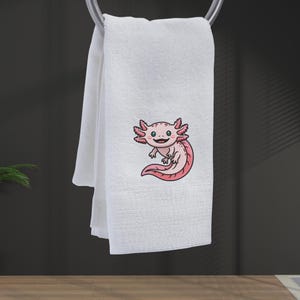May include: White hand towel with an embroidered cartoon axolotl in pink and red. The axolotl has a smiling face and is positioned on the lower right side of the towel. The towel is hanging from a silver towel rack.