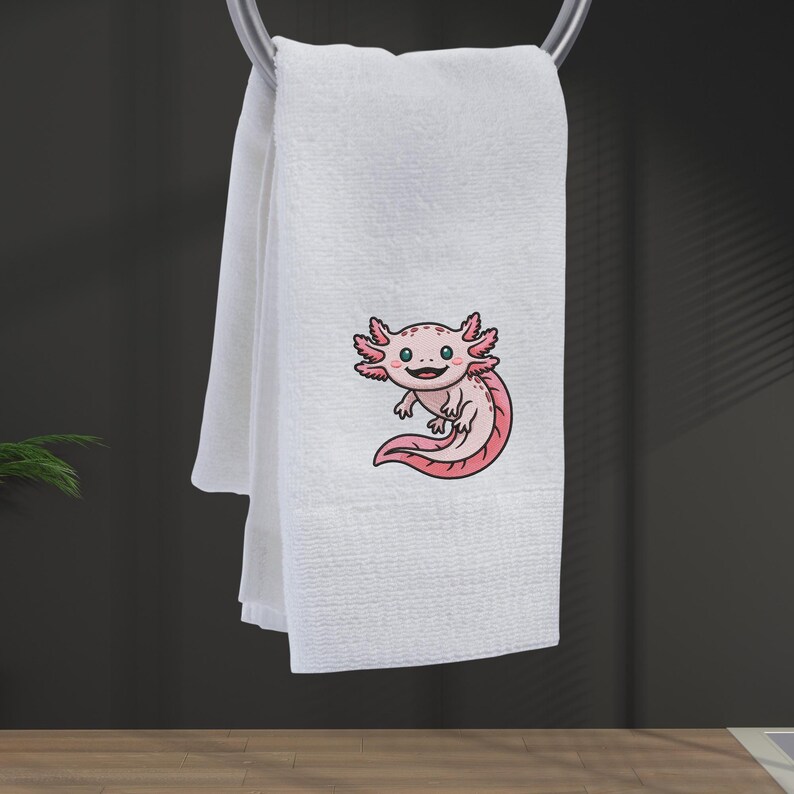 May include: White hand towel with an embroidered cartoon axolotl in pink and red. The axolotl has a smiling face and is positioned on the lower right side of the towel. The towel is hanging from a silver towel rack.