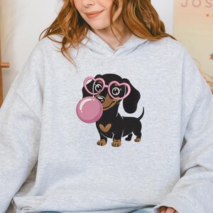 May include: A light gray hoodie featuring a cartoon dachshund wearing pink heart-shaped glasses and blowing a pink bubble. The dachshund is black and brown with a heart-shaped patch on its chest.
