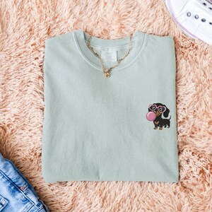 May include: A light green t-shirt featuring an embroidered dachshund wearing pink heart-shaped glasses and blowing a pink bubble. A gold necklace is draped on the shirt. The shirt is folded on a pink fluffy surface.