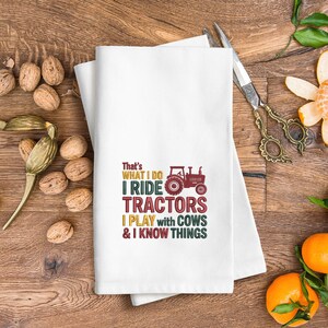 Peut inclure: Serviette de cuisine blanche avec le texte "That's WHAT I DO I RIDE TRACTORS I PLAY with COWS & I KNOW THINGS" en plusieurs couleurs. Un dessin de tracteur rouge est &eacute;galement inclus. La serviette est pli&eacute;e sur une surface en bois.