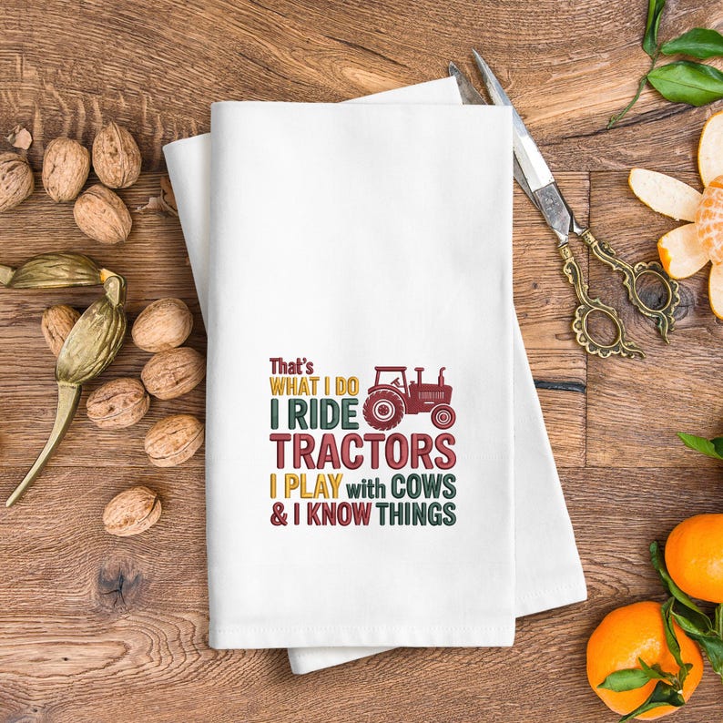 Peut inclure: Serviette de cuisine blanche avec le texte "That's WHAT I DO I RIDE TRACTORS I PLAY with COWS & I KNOW THINGS" en plusieurs couleurs. Un dessin de tracteur rouge est &eacute;galement inclus. La serviette est pli&eacute;e sur une surface en bois.