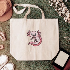 May include: A cream-colored tote bag featuring a cartoon axolotl with pink gills and tail. The axolotl has a smiling expression. The bag has two handles and is sitting on a green surface.