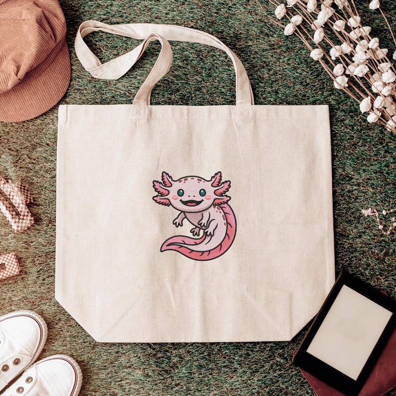 May include: A cream-colored tote bag featuring a cartoon axolotl with pink gills and tail. The axolotl has a smiling expression. The bag has two handles and is sitting on a green surface.