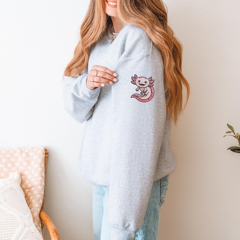 May include: A light gray sweatshirt featuring a pink axolotl graphic on the upper left sleeve. The axolotl has a smiling face and a pink body. The sweatshirt is a casual style.