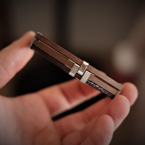 May include: A brown leather bracelet with silver-colored metal accents. The bracelet features multiple layers of leather in varying textures and shades of brown. A stylish accessory for everyday wear.