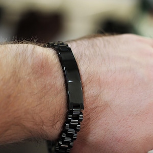 Może przedstawiać: Biały ceramiczny kubek z czarnym uchwytem i napisem "But First, Coffee" pogrubioną, czarną czcionką. Kubek jest ustawiony na rozmytym tle, sugerującym przytulne otoczenie.