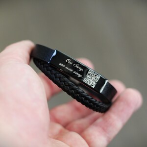 May include: Black bracelet with a braided leather band and a rectangular metal plate. The plate has the engraved text "Our Story, one scan away..." and a QR code. The bracelet is held in a hand.