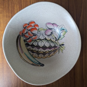 May include: A cream-coloured ceramic plate with a hand-painted fruit design. The design includes a banana, pineapple, and grapes in shades of yellow, orange, purple, and green. The plate has an irregular, organic shape.