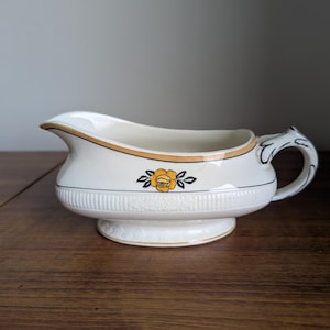 May include: Cream-coloured gravy boat with a decorative yellow rose design. The boat has a gold trim and a handle with a black and white design. The base is round and has a decorative pattern.