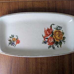 Könnte beinhalten: Rechteckige weiße Keramikservierplatte mit goldenem Rand und Blumendesigns. Die Platte hat zwei Blumencluster in Orange, Gelb und Lila. Ein Vintage-Stück zum Servieren von Vorspeisen oder zum Präsentieren kleiner Gegenstände.