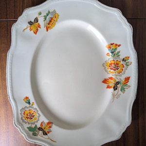 May include: Cream-coloured, scalloped-edge plate with floral designs in orange, yellow, and grey. The plate has a slightly oval centre and a decorative, metallic trim around the edge. A vintage-style serving dish.