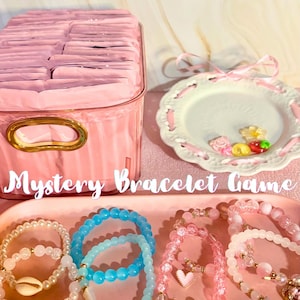 Può includere: Un contenitore in plastica rosa riempito con piccoli pacchetti rosa, un piatto decorativo bianco con nastro rosa e piccoli ciondoli, e un vassoio rosa contenente diversi braccialetti di perline con ciondoli, testo Mystery Bracelet Game.