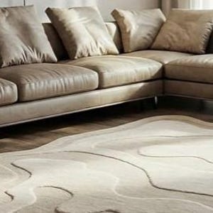 Pu&ograve; includere: Un divano angolare beige con diversi cuscini &egrave; posizionato sopra un tappeto chiaro con un motivo topografico ondulato. Il tappeto ha un bordo irregolare e il divano ha un design moderno e minimalista.
