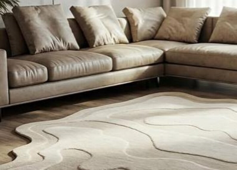 Pu&ograve; includere: Un divano angolare beige con diversi cuscini &egrave; posizionato sopra un tappeto chiaro con un motivo topografico ondulato. Il tappeto ha un bordo irregolare e il divano ha un design moderno e minimalista.